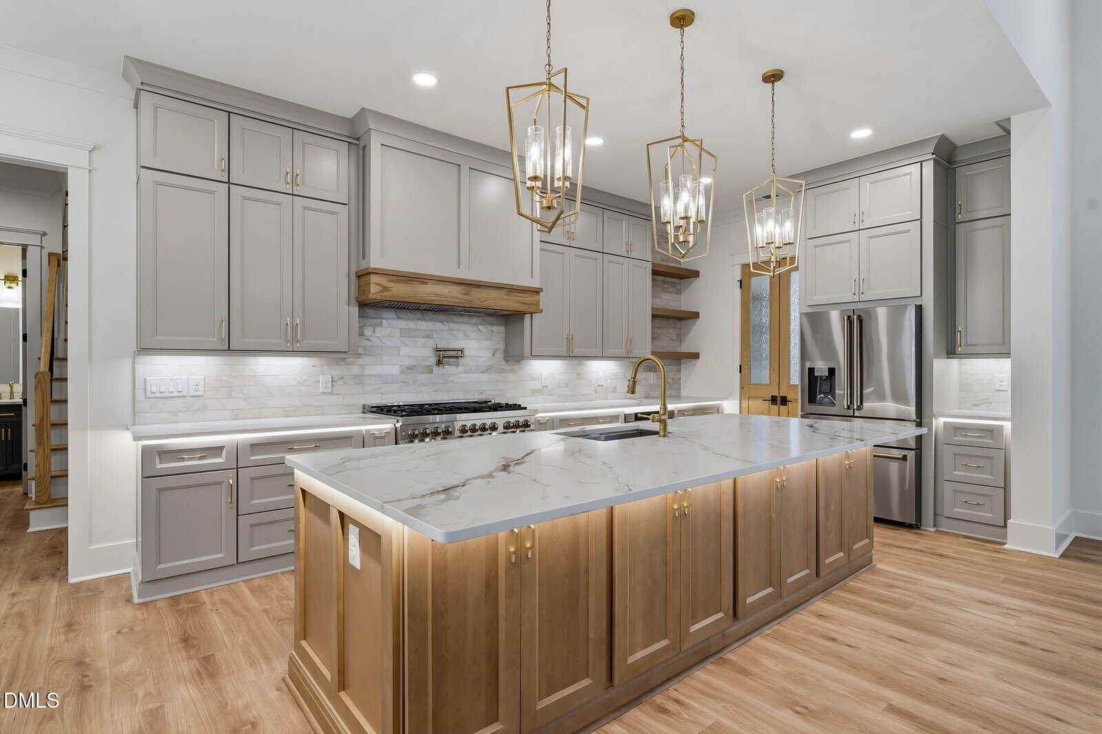 Modern kitchen with light gray cabinets, a large marble island with wood base, stainless steel appliances, gold pendant lights, and wood flooring. Marble backsplash and under-cabinet lighting complete the clean, elegant look.