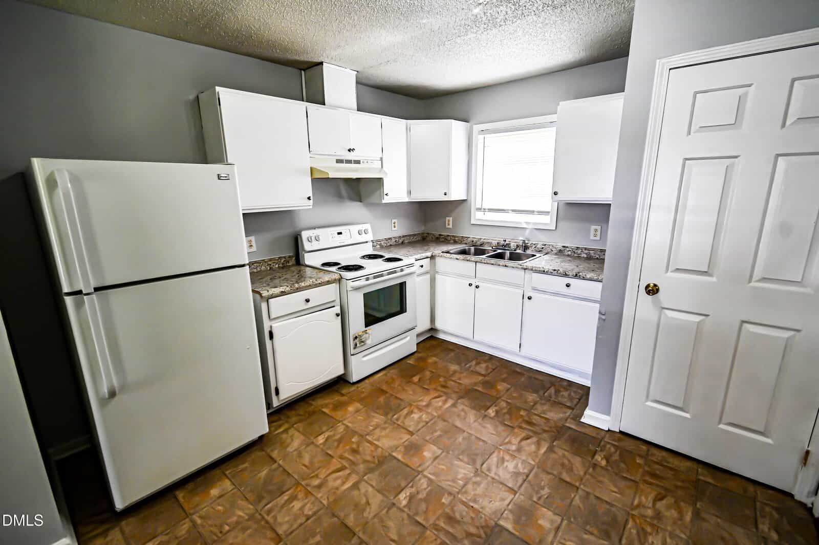 A kitchen with white cabinets, a white refrigerator, white stove, white range hood, double sink, brown patterned tile floor, and a window above the sink. Walls are painted gray and a white door is visible on the right.