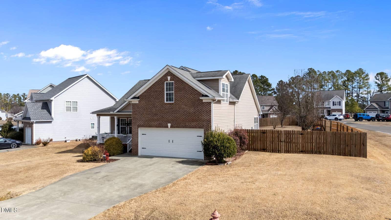 A two-story suburban house with a brick and siding exterior, white garage door, and concrete driveway. The yard is dry with minimal landscaping, and a wooden fence encloses the backyard. Neighboring houses are visible.