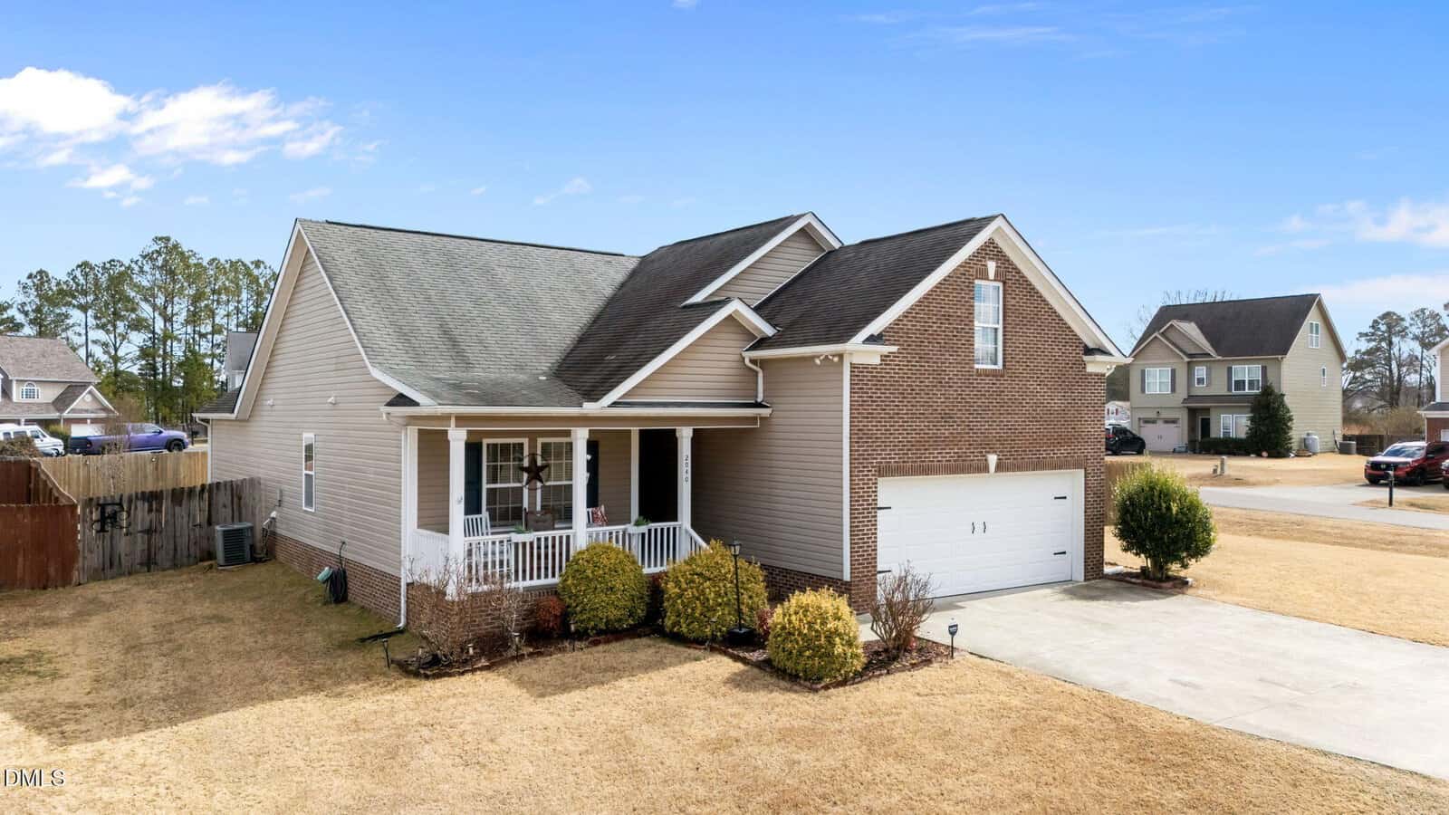 A single-story suburban house with tan siding, partial brick facade, white trim, and a covered front porch. The home features a two-car garage and a driveway. The lawn is mostly dry grass with several small bushes.