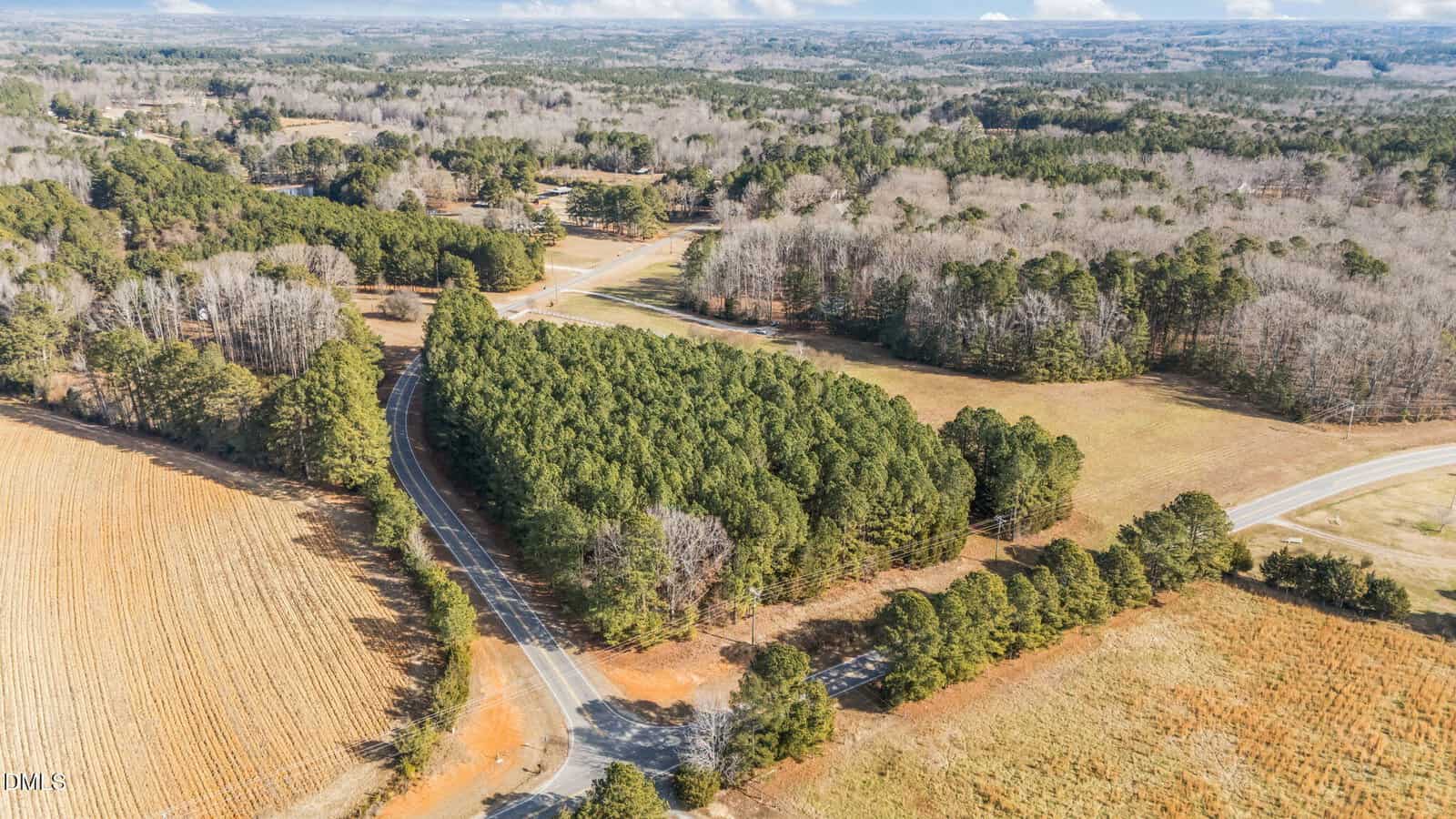 Aerial view of a rural landscape with a winding road passing through wooded areas and open fields, bordered by trees and patches of farmland under a partly cloudy sky.