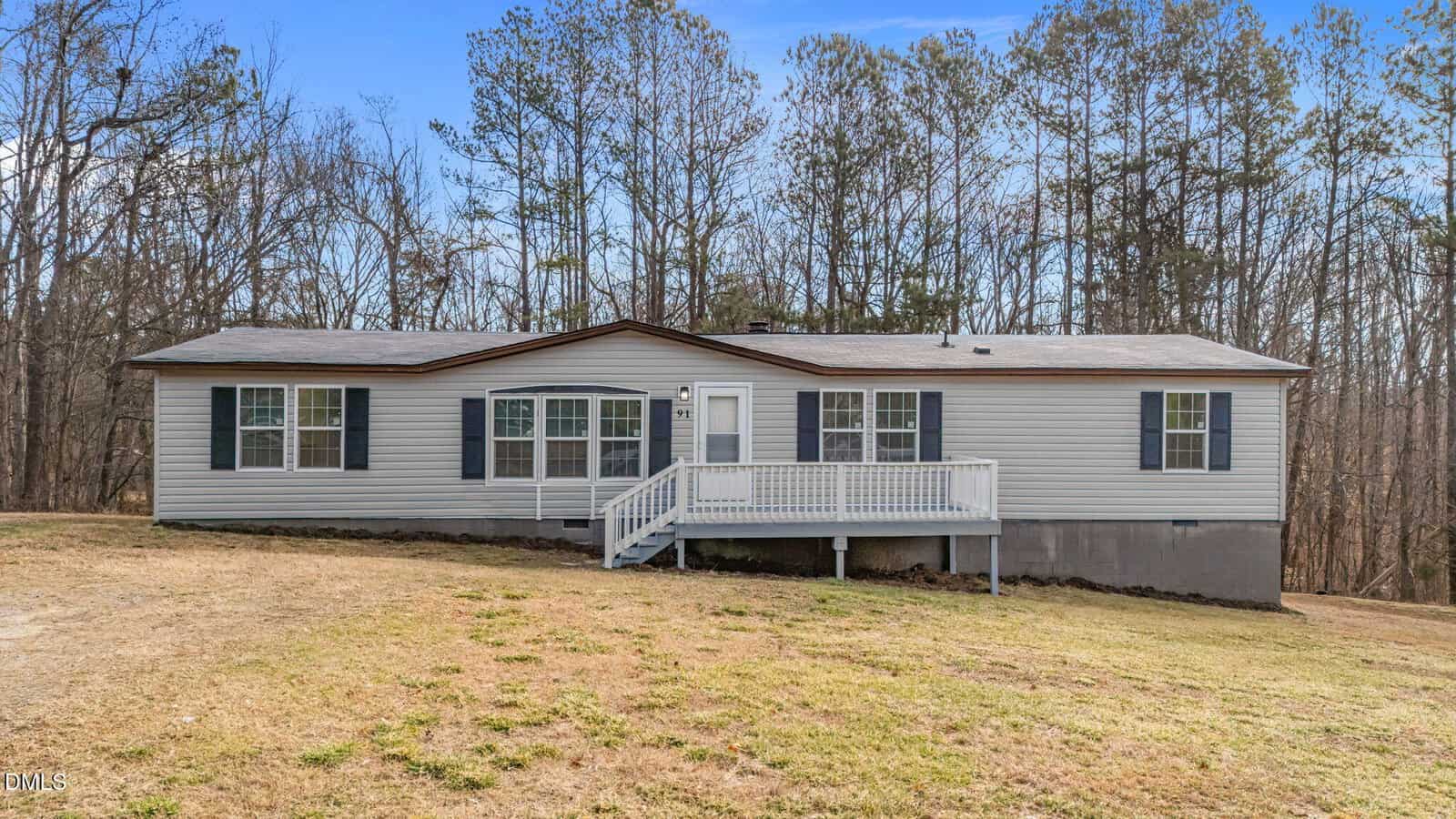 Single-story manufactured home at 91 Pine Ridge Trail in Henderson, featuring gray siding, black shutters, and a small front porch with white railing. The house sits on a sloped grassy yard bordered by trees with no leaves under a clear blue sky.