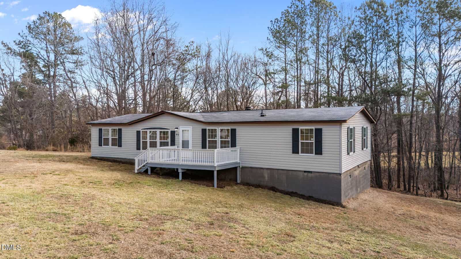 Single-story manufactured home with gray siding, black shutters, and a small front porch. The house is set on a sloped grassy yard, bordered by leafless trees and evergreens under a partly cloudy sky.