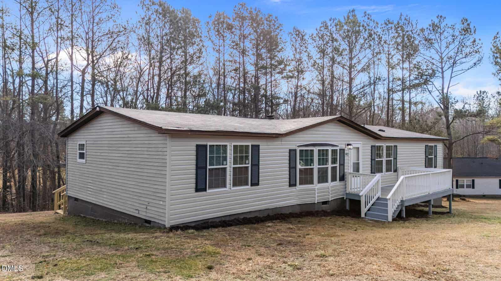 Single-story beige manufactured home with dark trim, multiple windows, a front porch with white railings, and steps. The house sits on a sloped grassy yard, surrounded by tall trees and another home in the background.