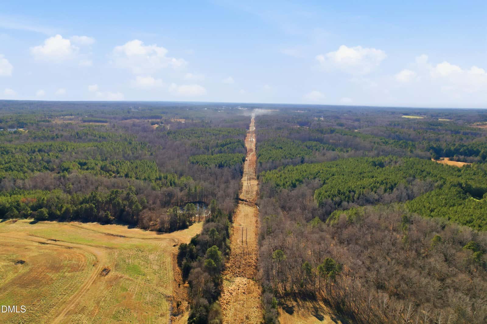 Aerial view of a long, cleared utility corridor cutting through dense forest and fields under a bright blue sky with scattered clouds.