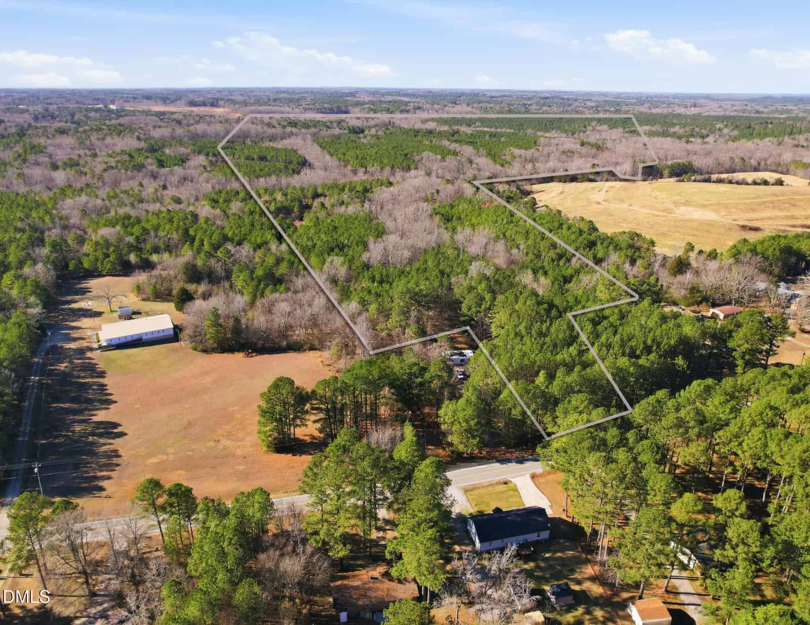 Aerial view of a rural, wooded landscape with outlined property boundaries, open fields, clusters of trees, a few buildings, and a small road winding through the area under a partly cloudy sky.