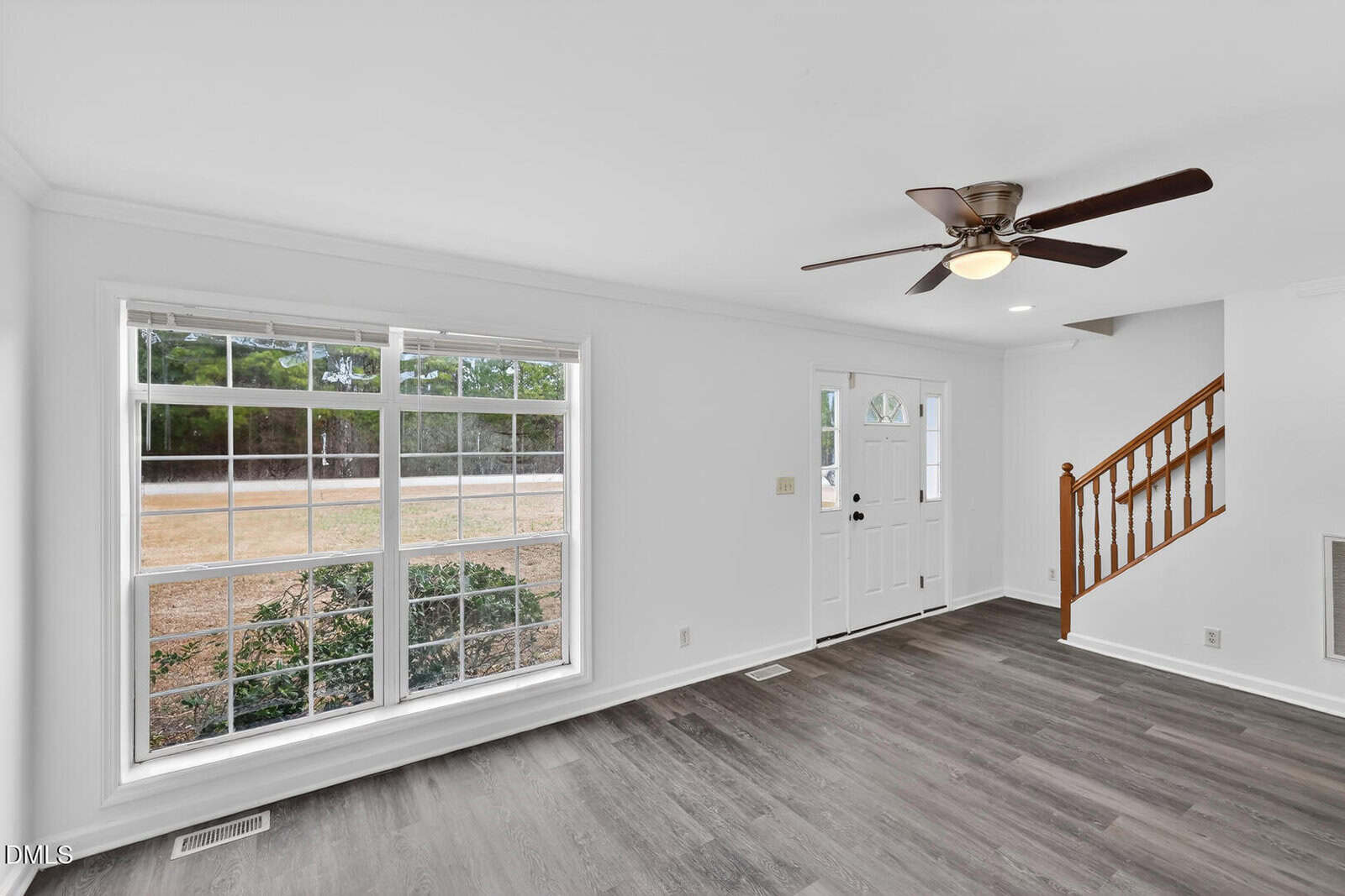 A bright living room with large windows, a white front door with glass panels, dark wood flooring, white walls, and a wooden staircase with a banister on the right side. A ceiling fan with lights is mounted above.