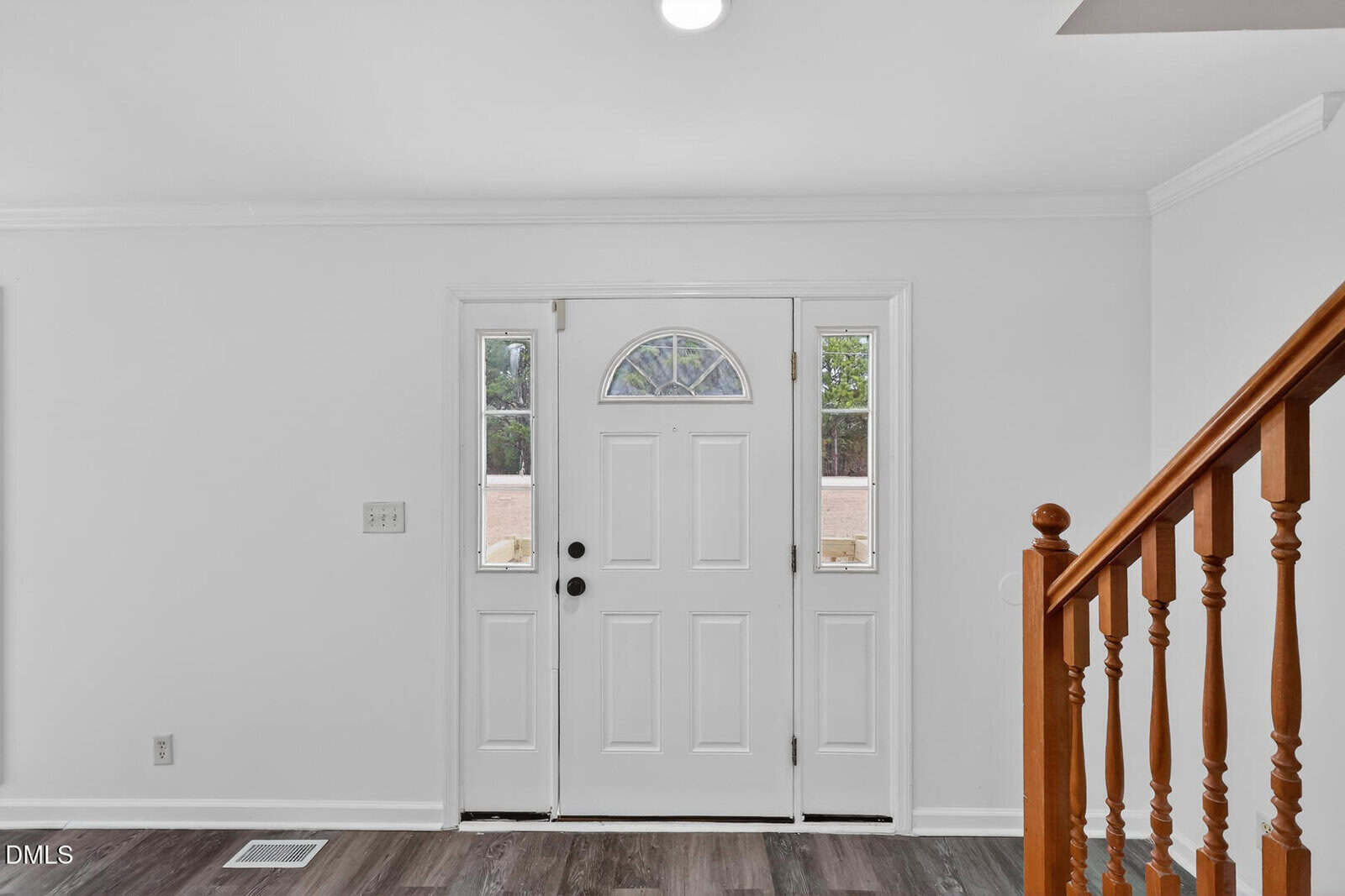 A white front door with a semi-circular window at the top is centered in an entryway. The floor is dark wood, and a wooden staircase with spindles is visible on the right side. Walls are painted white.