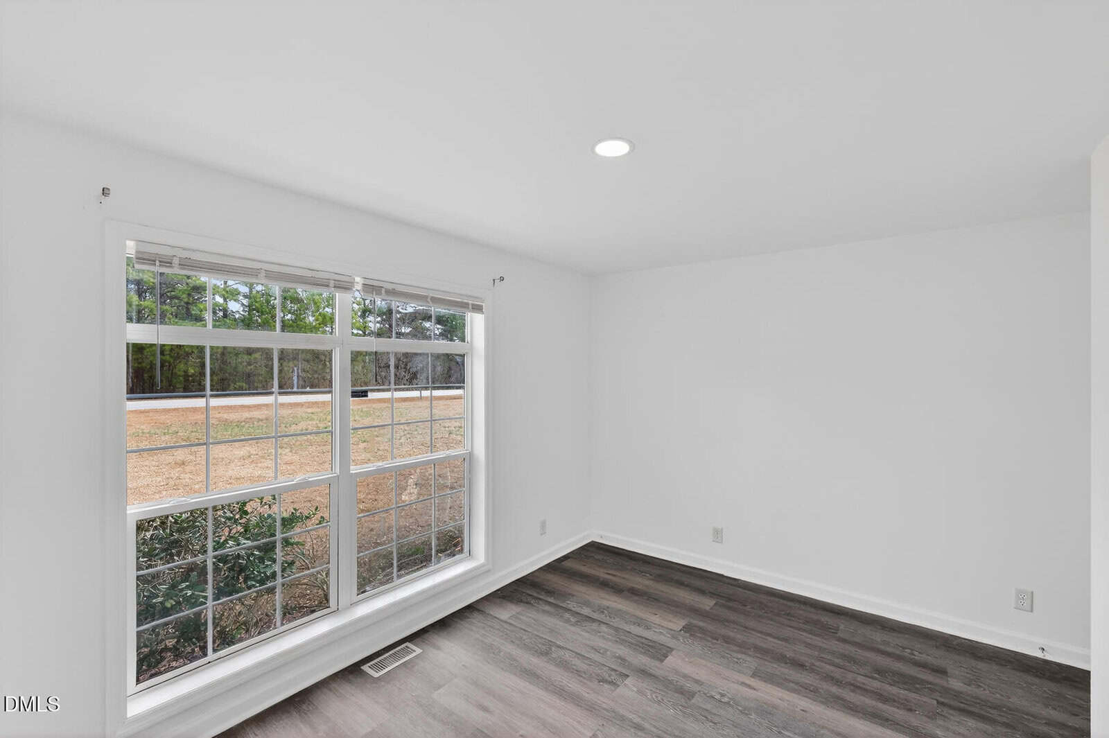 A bright, empty room with white walls, a large window with grid panes, and dark wood flooring. The window overlooks a yard with grass and trees. A single recessed ceiling light is visible.