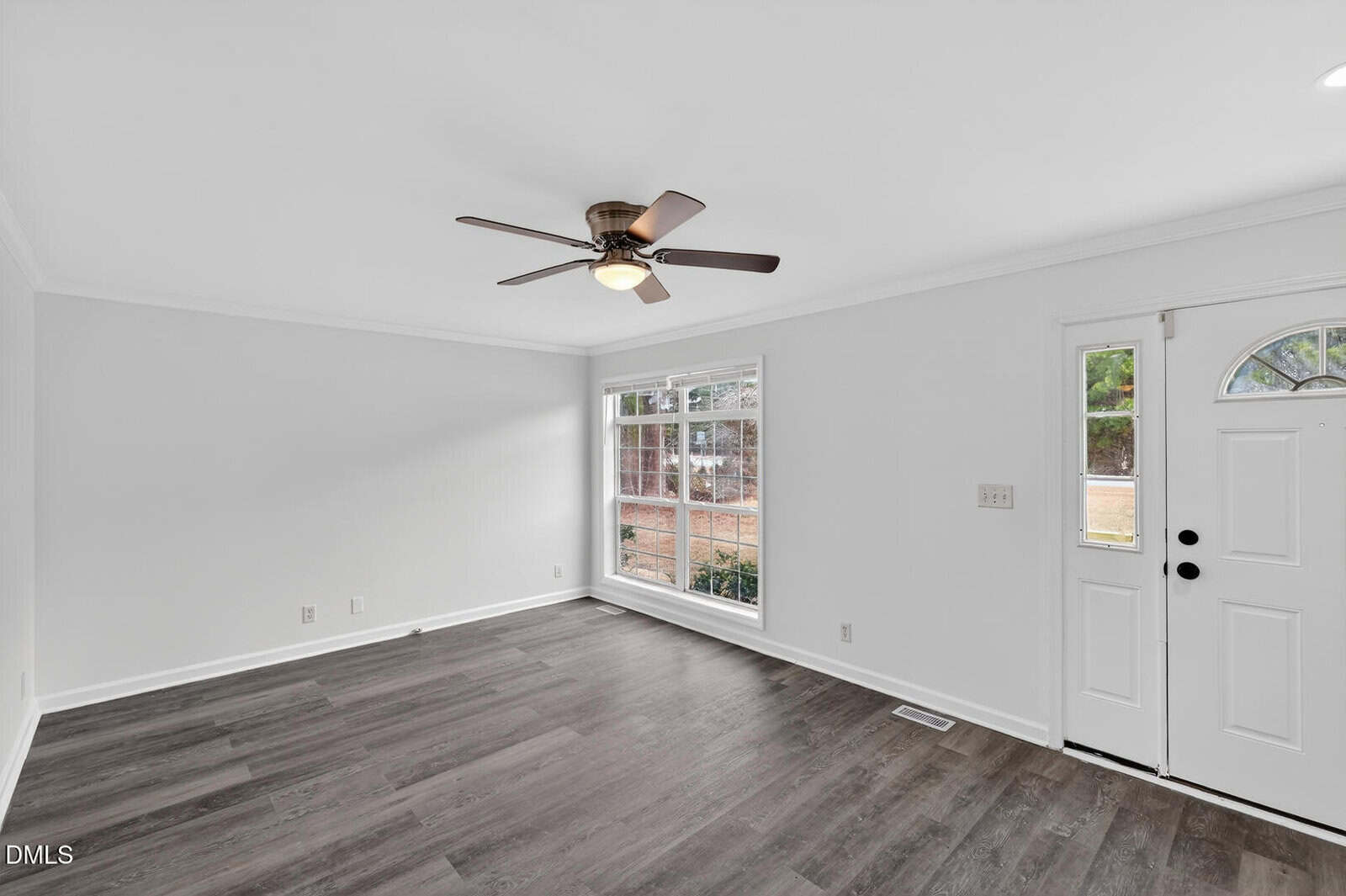 A bright, empty room with white walls, dark wood flooring, a ceiling fan with light, a large window letting in natural light, and a white front door with a small window.