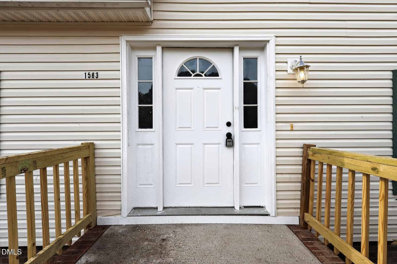 A white front door with a semicircular window, flanked by two narrow windows. The house has beige siding, a brass lantern-style light, two wooden ramps, and the number 1583 displayed to the left of the door.