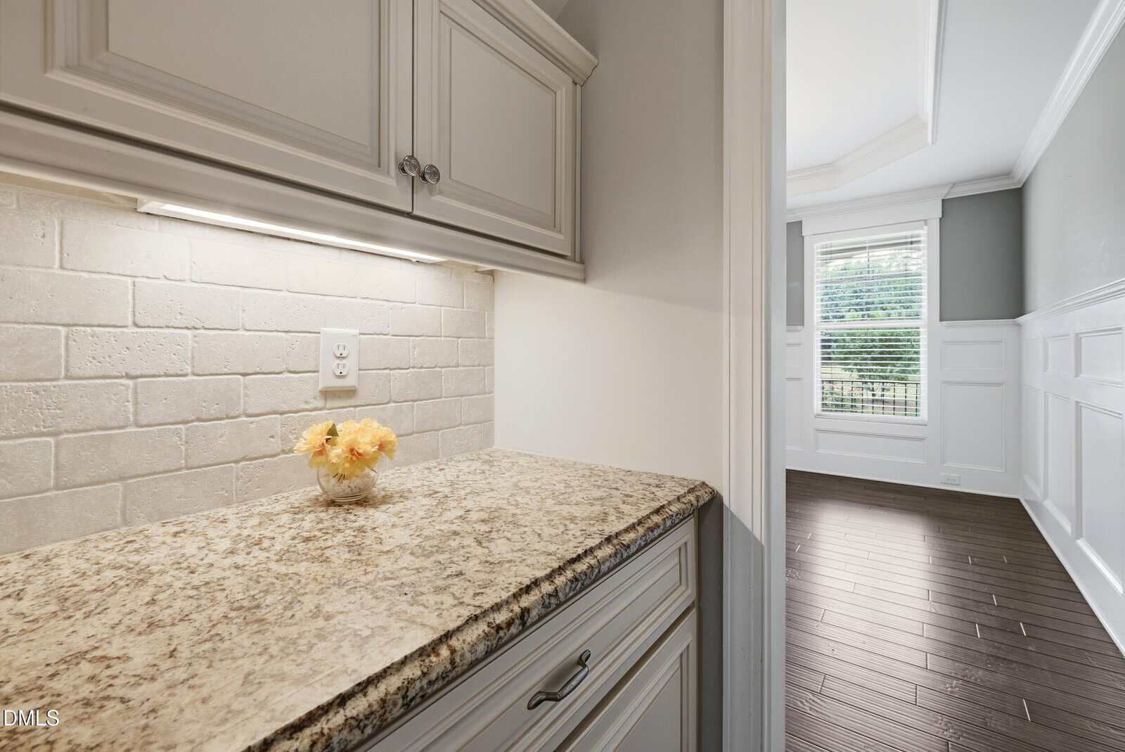 A small granite countertop with white cabinets and a tile backsplash, featuring a vase of yellow flowers. The area opens to a room with dark wood flooring, wainscoting, and a window with blinds.