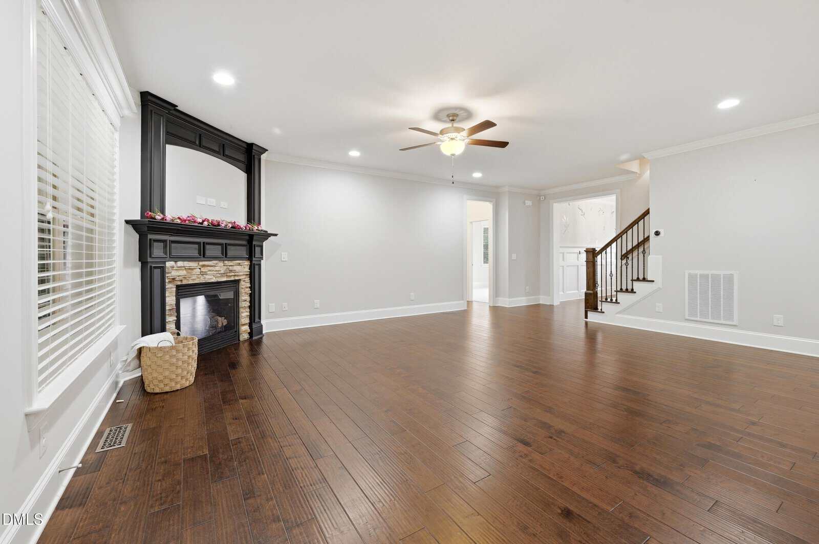 Spacious living room with hardwood floors, white walls, a ceiling fan, recessed lights, a black fireplace with a stone surround, large windows with blinds, and a wooden staircase leading upstairs.