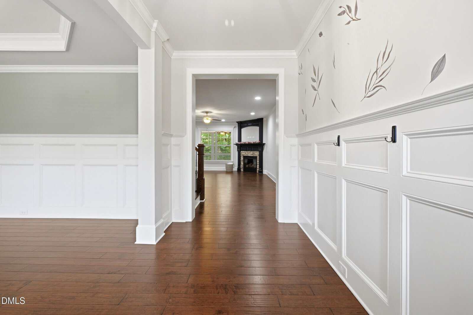 A hallway with white wainscoting, light walls with leaf designs, dark wood floors, and hooks on the right wall, leading to a living area with a fireplace and large windows.