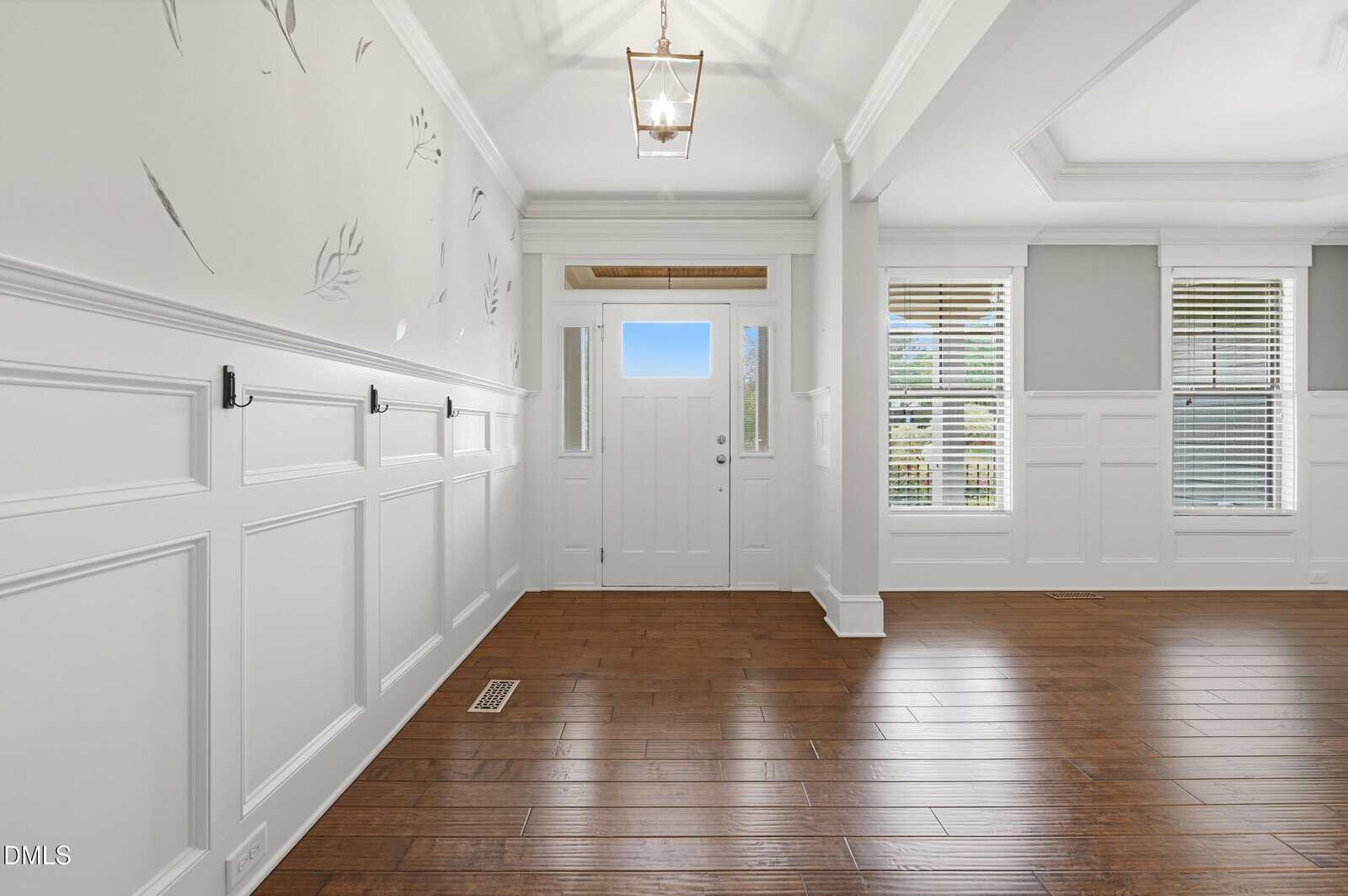 Bright entryway with white paneled walls, hooks, and hardwood floors; a white front door with glass panels lets in light, and large windows are visible in the adjacent room.