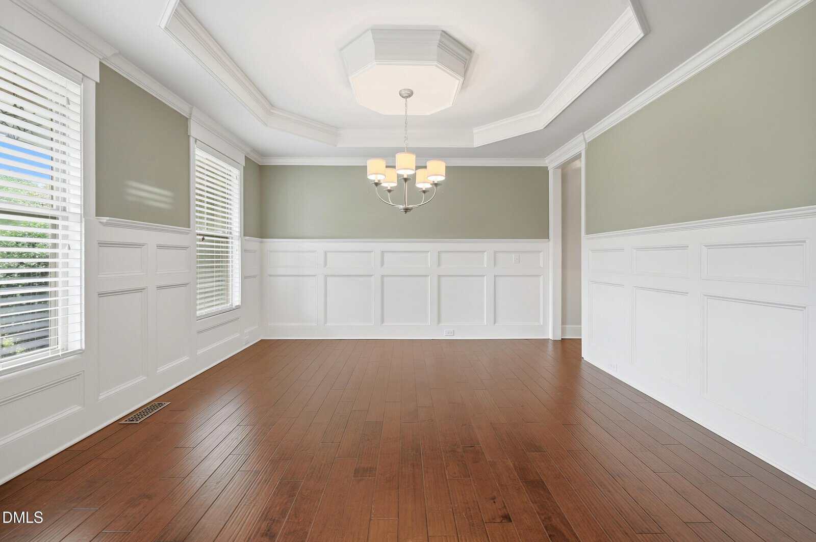 Empty dining room with light green upper walls, white wainscoting, and medium brown wood floors. Three windows with white blinds and a ceiling light fixture are present. There is natural light coming in from the left.