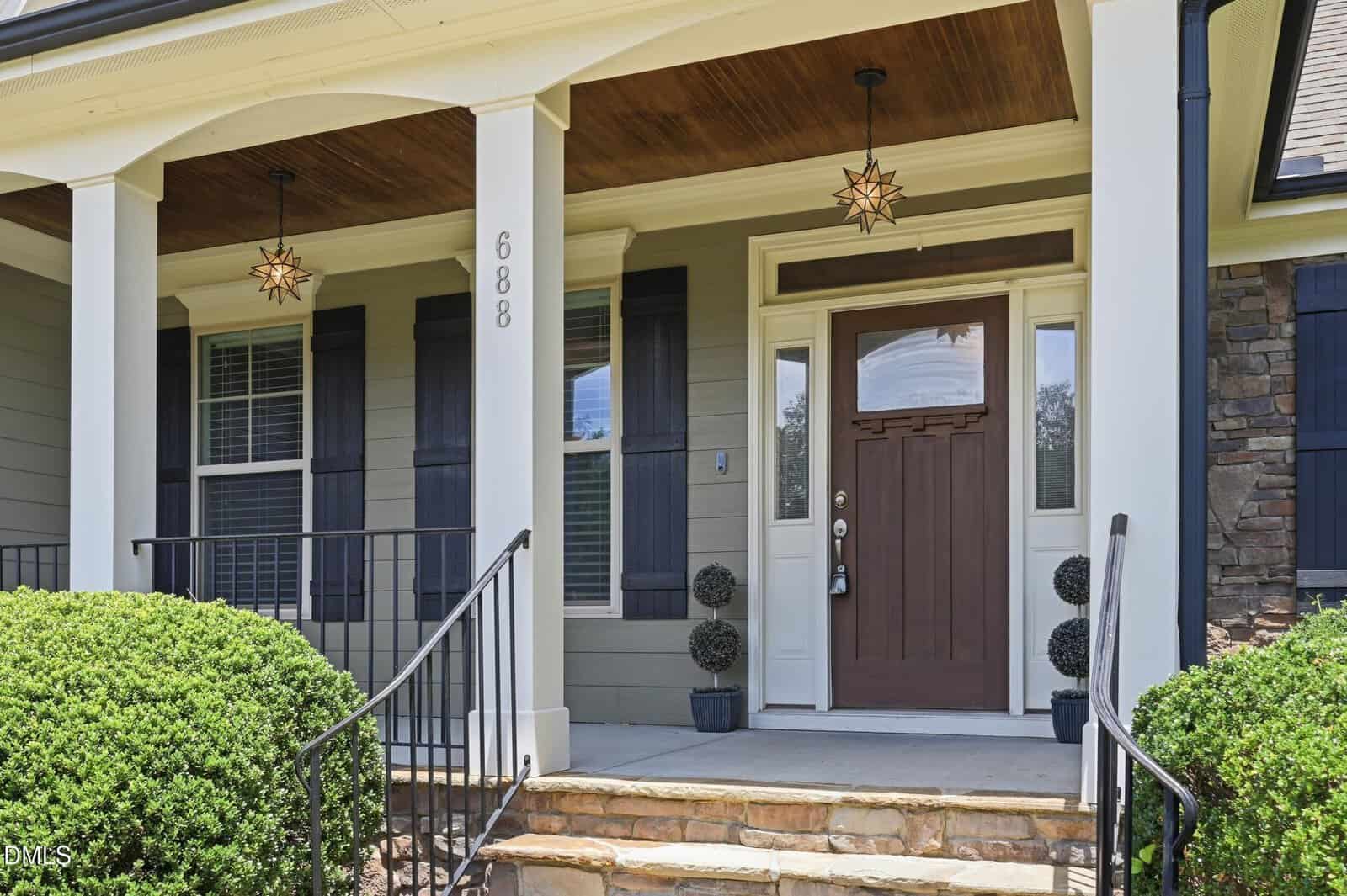 A house front porch with a brown door, white trim, gray siding, two star-shaped pendant lights, black railings, potted topiary plants, and stone steps surrounded by green shrubs.