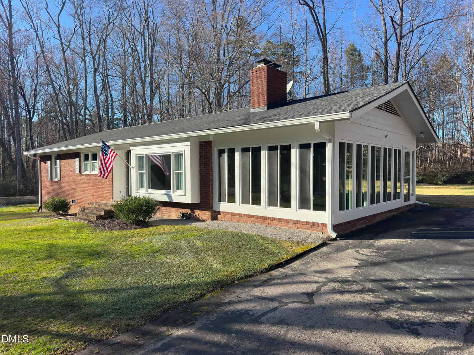 Single-story brick house with a large sunroom, American flag by the front door, a chimney, and surrounded by grass and leafless trees under a clear blue sky.