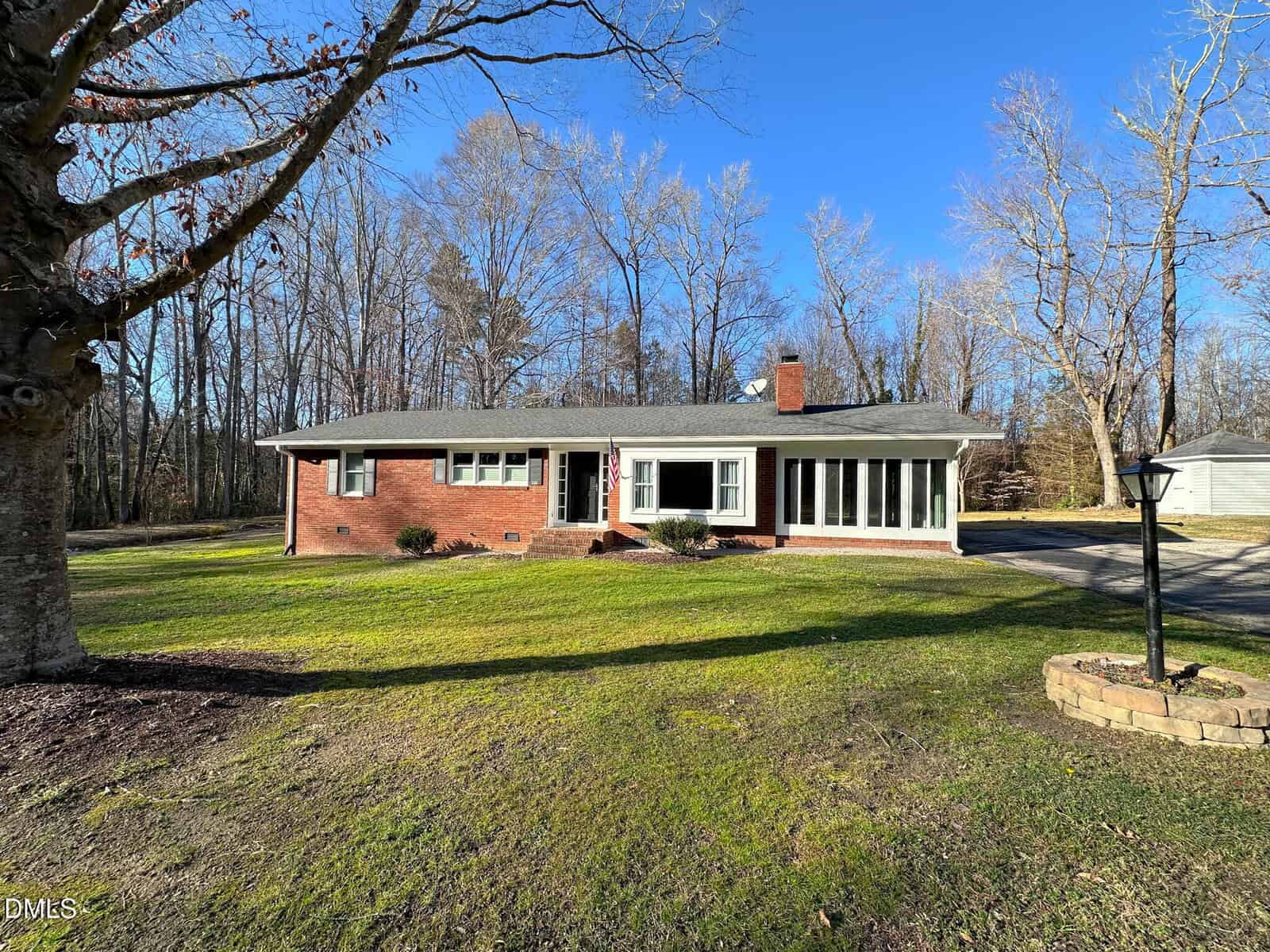 Single-story red brick house with large windows, a chimney, and a spacious front yard, surrounded by leafless trees under a clear blue sky. A driveway leads to a detached garage on the right.