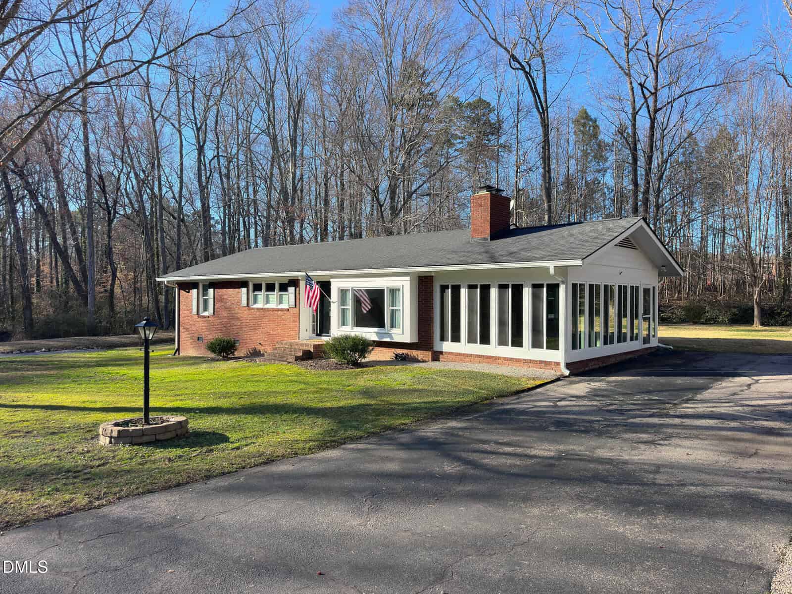 Charming single-story brick house at 330 Hazelwood Drive, Warrenton, featuring large front windows, an attached sunroom, and an American flag by the entrance. Surrounded by a well-kept lawn, mature trees, and a paved driveway, this home also boasts a small lamp post in the yard.