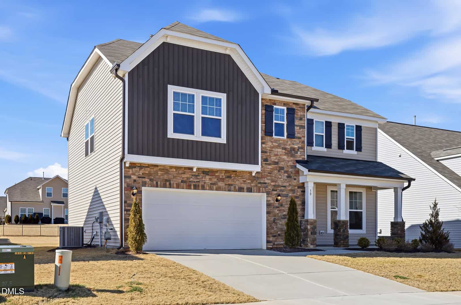 Two-story suburban house with a mix of stone, siding, and board-and-batten exterior, white garage door, paved driveway, and small bushes in front. The sky is clear and blue.