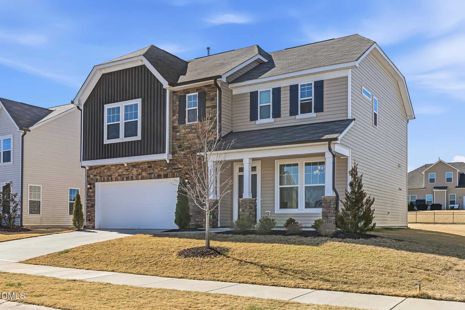 Two-story suburban house with beige siding, dark brown accent, and stone details. White-framed windows, a double garage, small front porch, and a leafless tree in the yard. The lawn is brown and recently mowed.