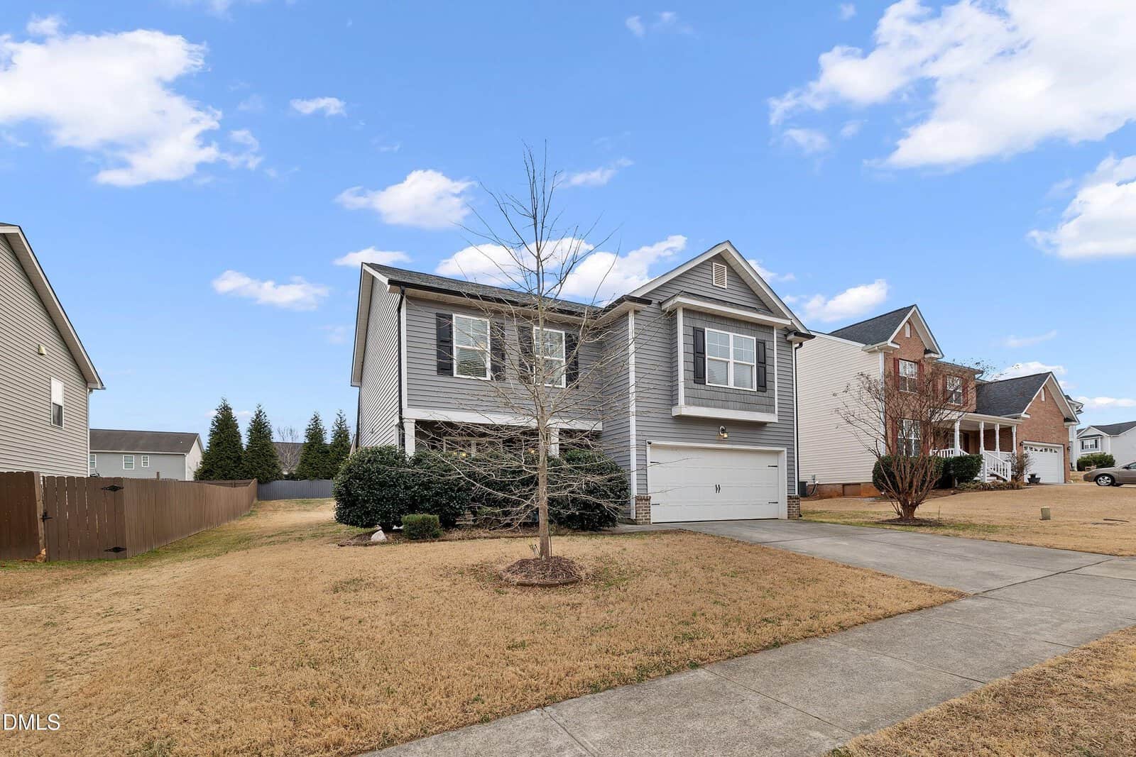 Two-story suburban house with gray siding, white trim, and a double garage. A leafless tree stands in the front yard, and the lawn appears dry. The house is surrounded by similar homes under a blue sky with scattered clouds.