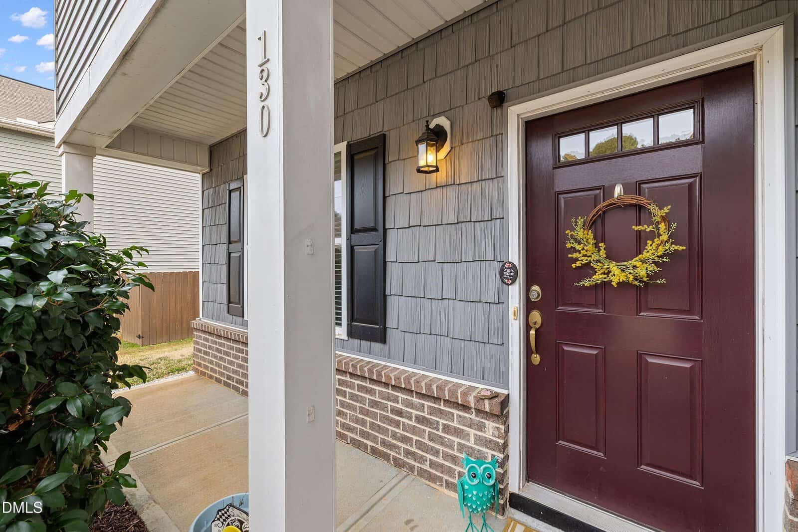 A front porch with gray siding, a maroon door decorated with a yellow floral wreath, a wall-mounted light, and an owl-shaped decoration on the ground near the step. House number 130 is visible on a white pillar.