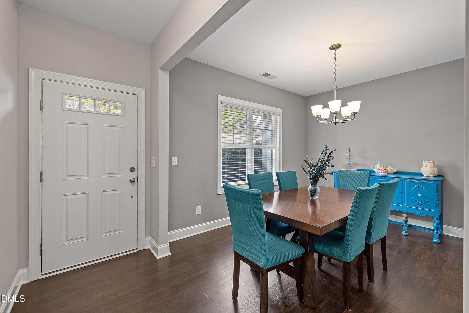 A modern dining room with a wooden table, six teal chairs, a bright blue sideboard, a vase centerpiece, and a chandelier. There is a white front door and large window letting in natural light.