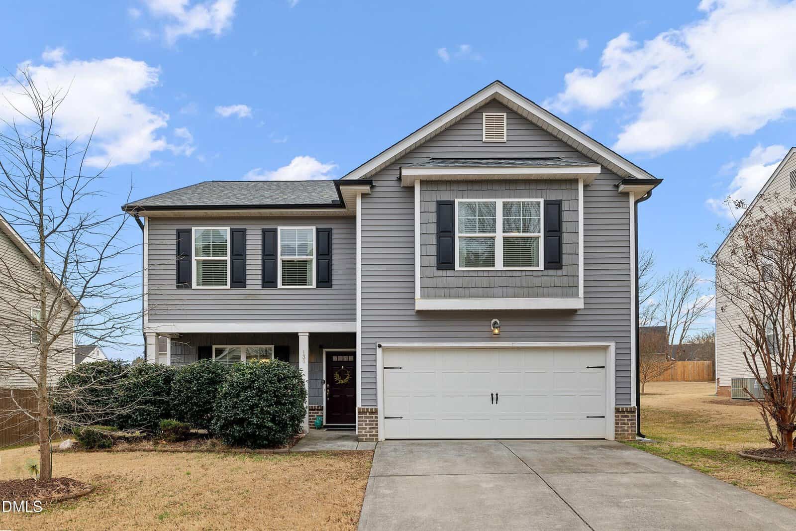 Two-story gray house with white trim, black shutters, and a double garage at 130 Ambergate Drive in Youngsville. The front yard features a driveway, some bushes, a bare tree, and a small lawn under a blue sky with a few clouds.