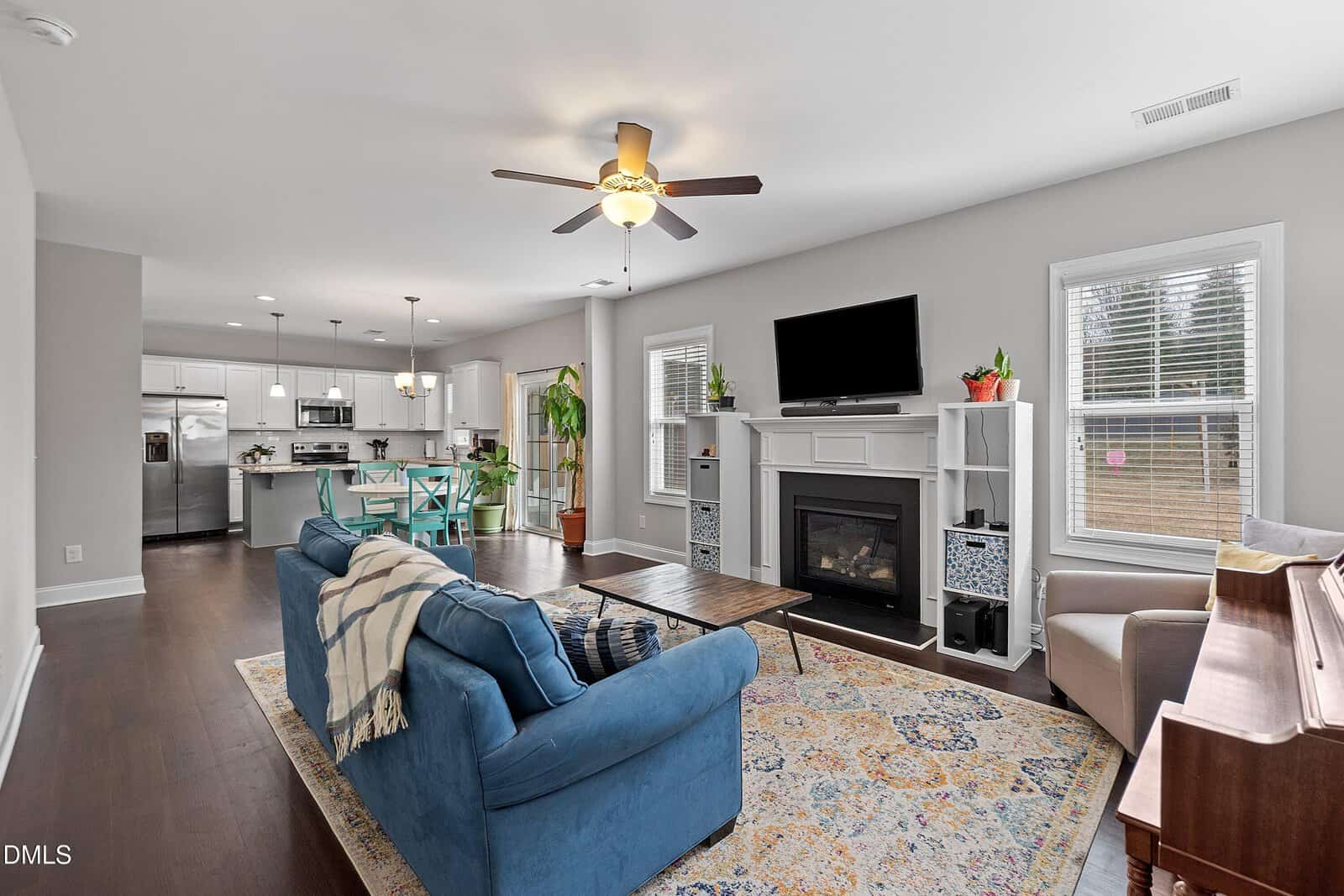 Open-concept living room with a blue sofa, wooden coffee table, patterned rug, fireplace, TV, piano, and adjacent dining area. Modern kitchen with white cabinets and stainless steel appliances in the background.