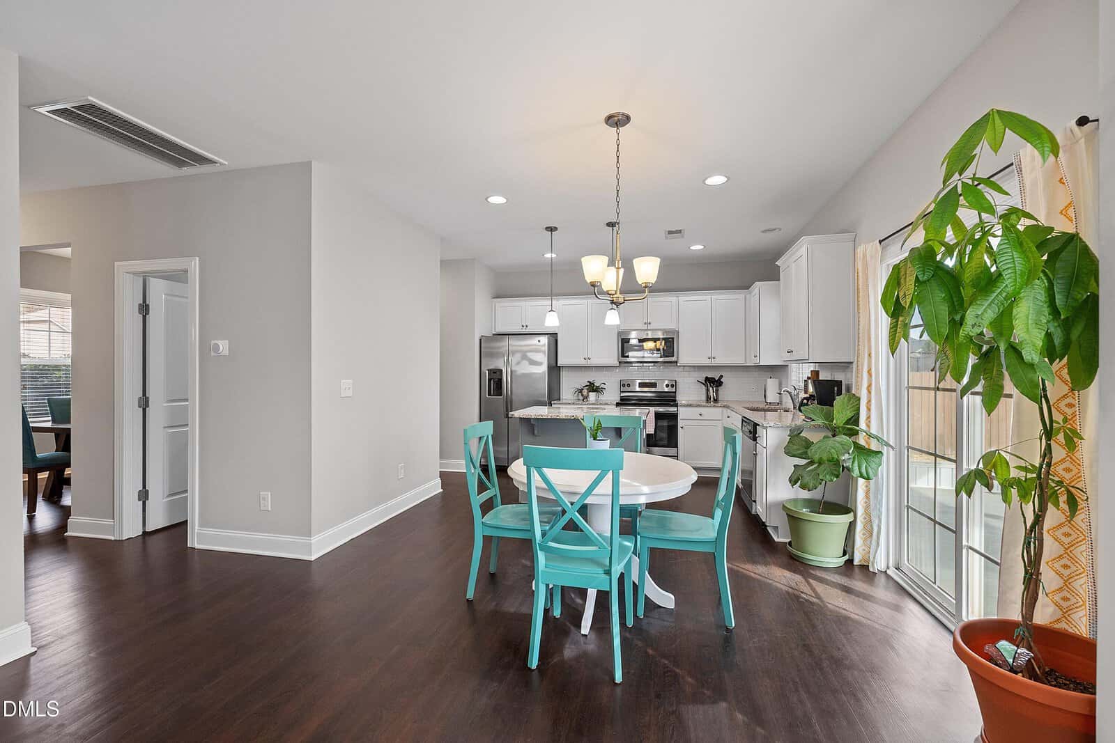 A modern kitchen and dining area with dark wood floors, white cabinets, stainless steel appliances, and a round table with four teal chairs. Large windows and glass doors let in natural light; several potted plants are present.
