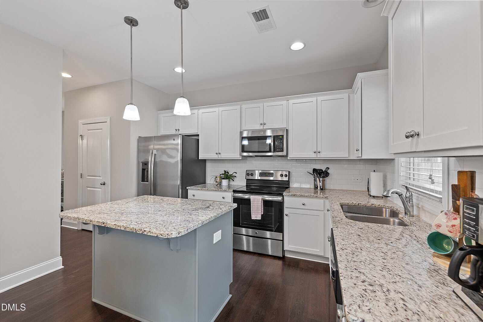 Modern kitchen with white cabinets, stainless steel appliances, granite countertops, a center island with pendant lights, dark wood flooring, and a large window above the sink. A towel hangs on the oven handle.