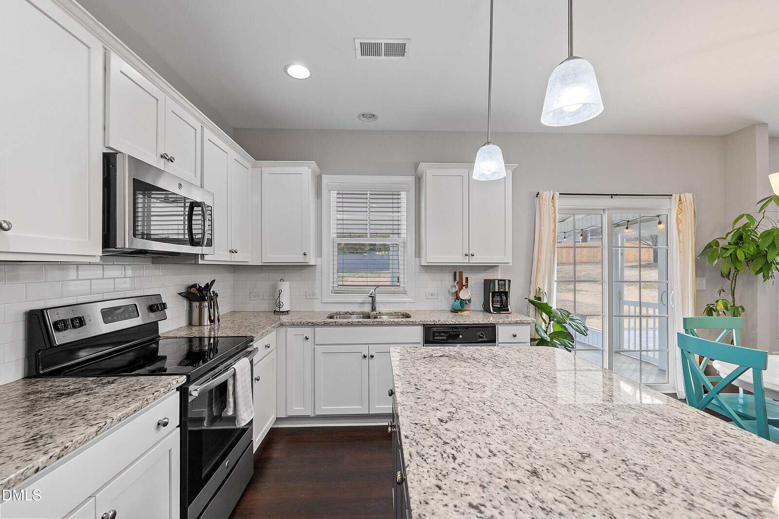 Modern kitchen with white cabinets, granite countertops, stainless steel appliances, a kitchen island, and pendant lights. A window is above the sink, and a dining area with a sliding glass door and plant is visible in the background.