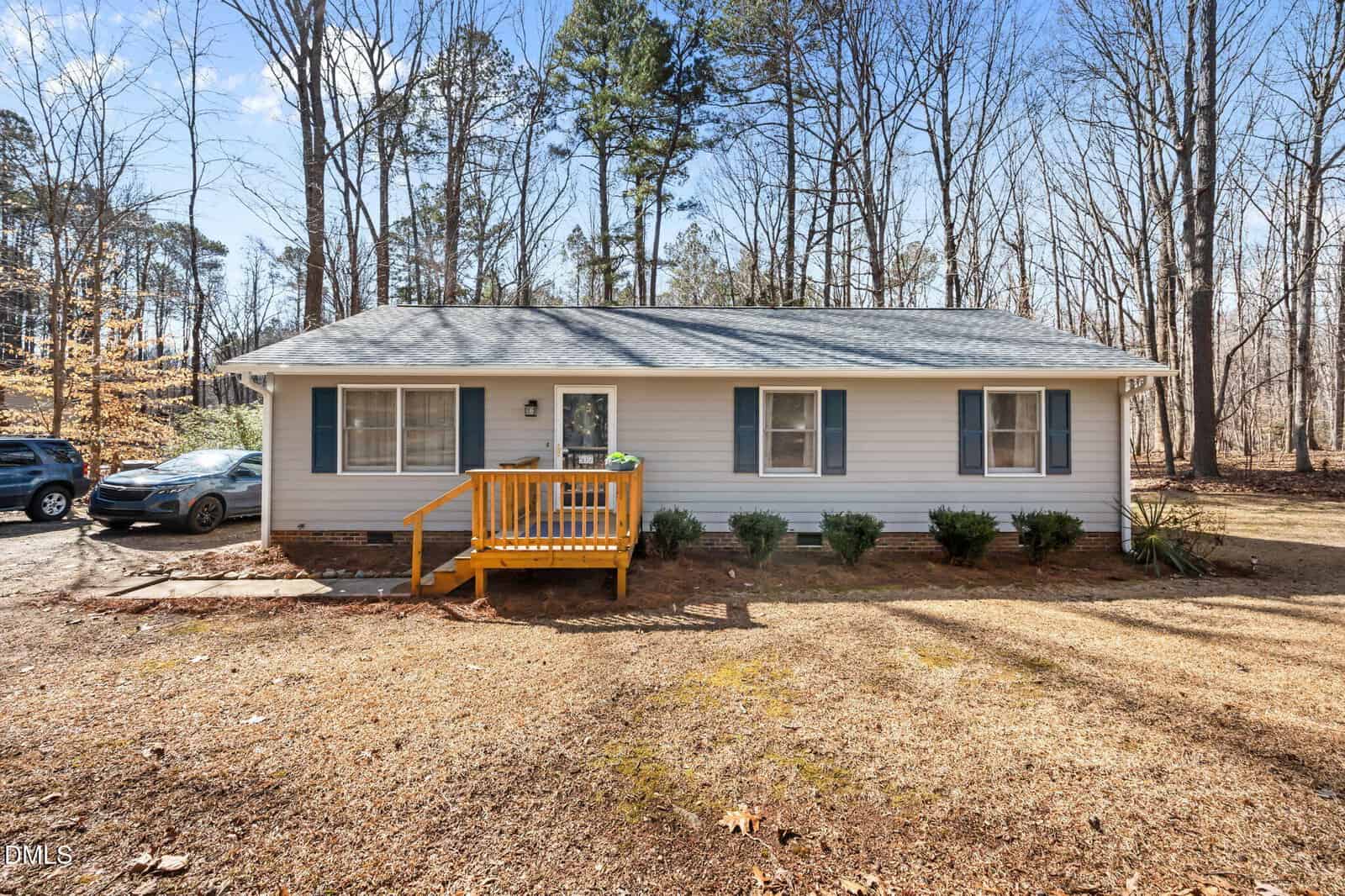 A single-story, light gray house with a small wooden front porch, blue shutters, and two front windows sits at 517 John Mitchell Road in Youngsville. The yard is mostly brown grass with some bushes, and trees surround the property. A black car is parked to the left.