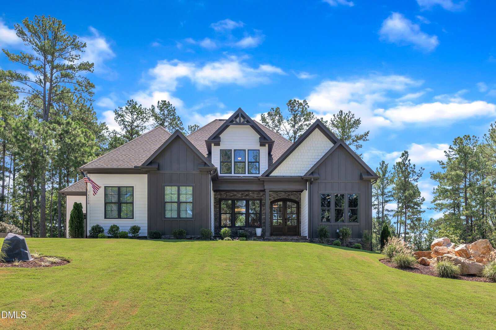 Nestled at 1120 Springdale Drive in Wake Forest, this two-story house features a striking blend of white and gray siding, a classic gabled roof, and large windows that fill the home with natural light. Set on a spacious green lawn dotted with trees and framed by a blue sky, the welcoming entrance proudly displays an American flag.