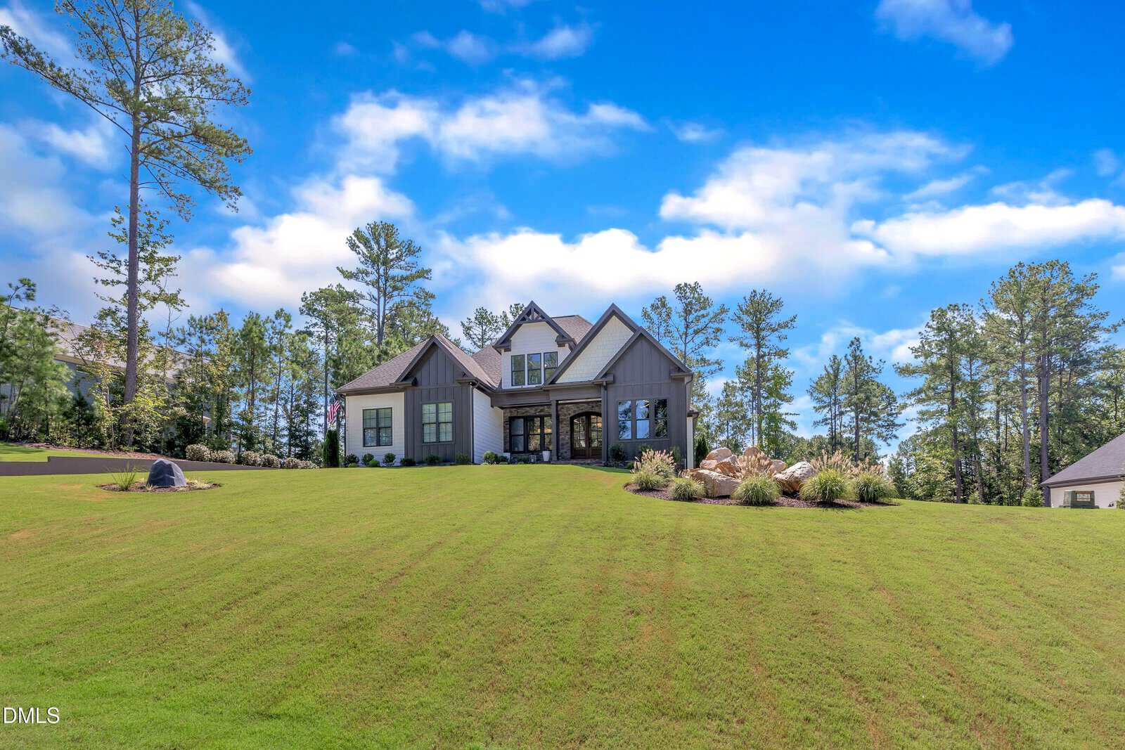 A two-story house with gray siding and stone accents sits on a large, neatly mowed lawn. Mature trees surround the property under a mostly blue sky with scattered clouds.