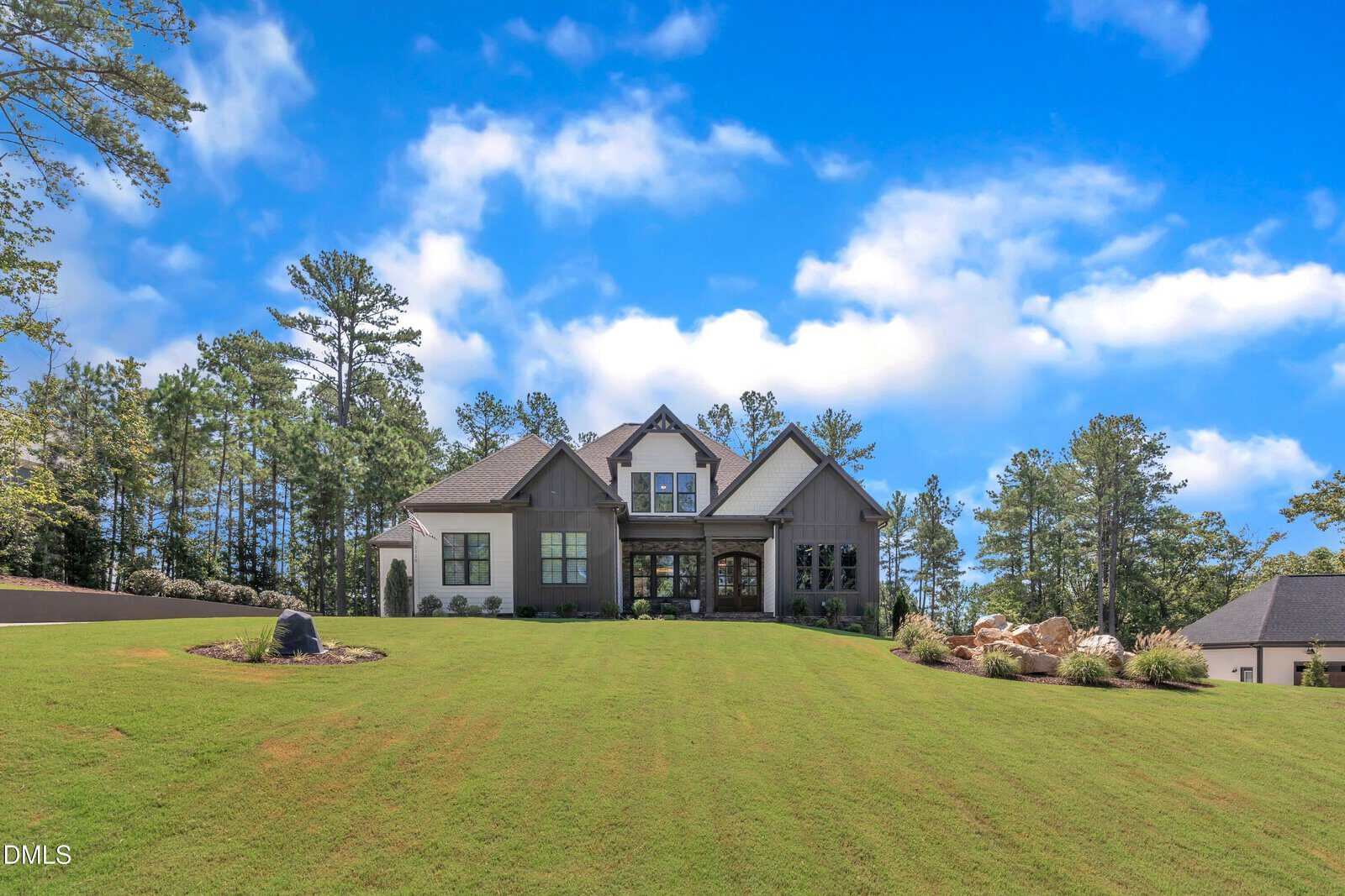 A large gray house with multiple gables sits on a wide, sloping green lawn, surrounded by trees under a blue sky with scattered clouds. Landscaping with rocks and bushes frames the front of the home.