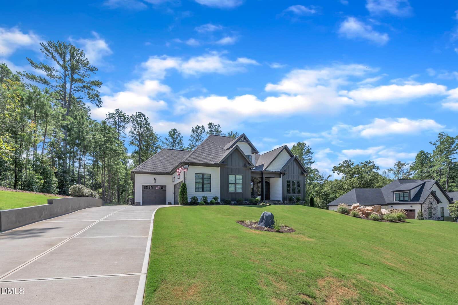 A modern single-story house with gray and white exterior, attached garage, and neatly landscaped lawn sits on a sloped hill under a blue sky with scattered clouds. A long concrete driveway leads up to the house.