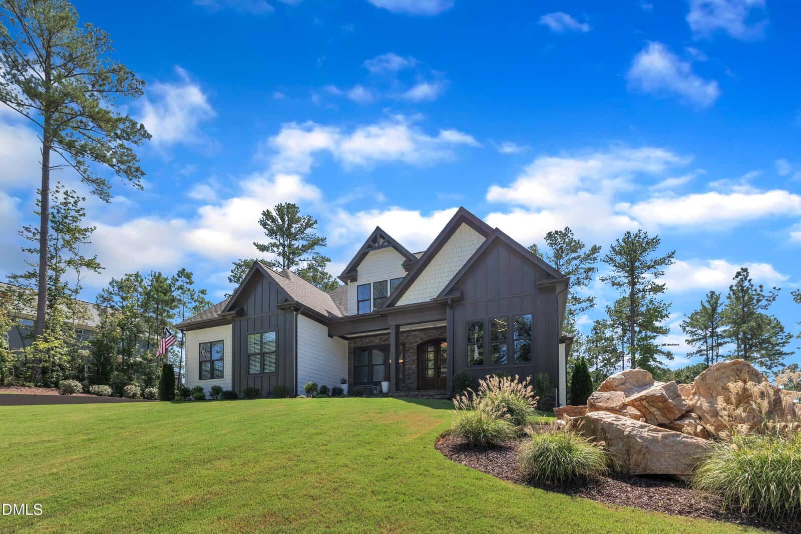 A modern two-story house with white siding and brown trim sits on a well-manicured lawn with landscaping rocks, shrubs, and trees under a bright blue sky with scattered clouds.