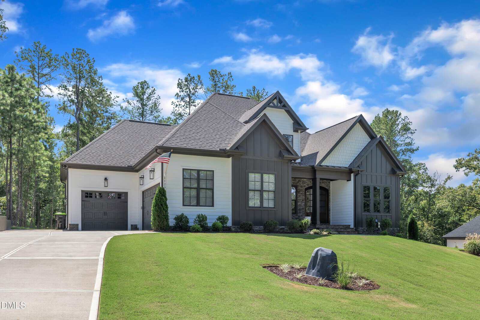 Modern two-story house with a gray roof, front porch, attached garage, large windows, and landscaped yard on a sloped lawn with a driveway and American flag by the entrance. Trees and blue sky are in the background.