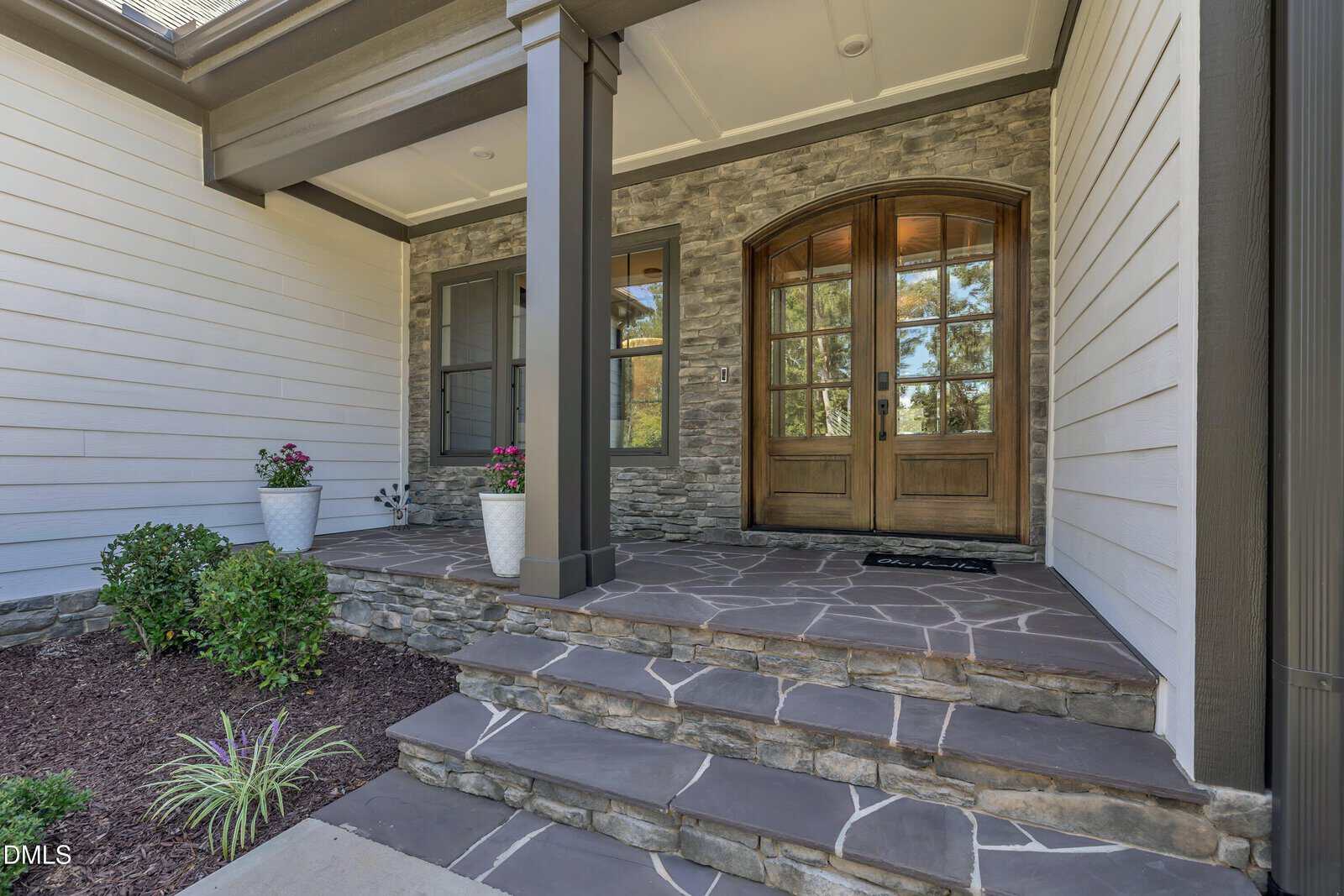 Stone steps lead to a covered entryway with double wooden doors, large windows, potted plants, and stone and wood siding on the exterior of a house.