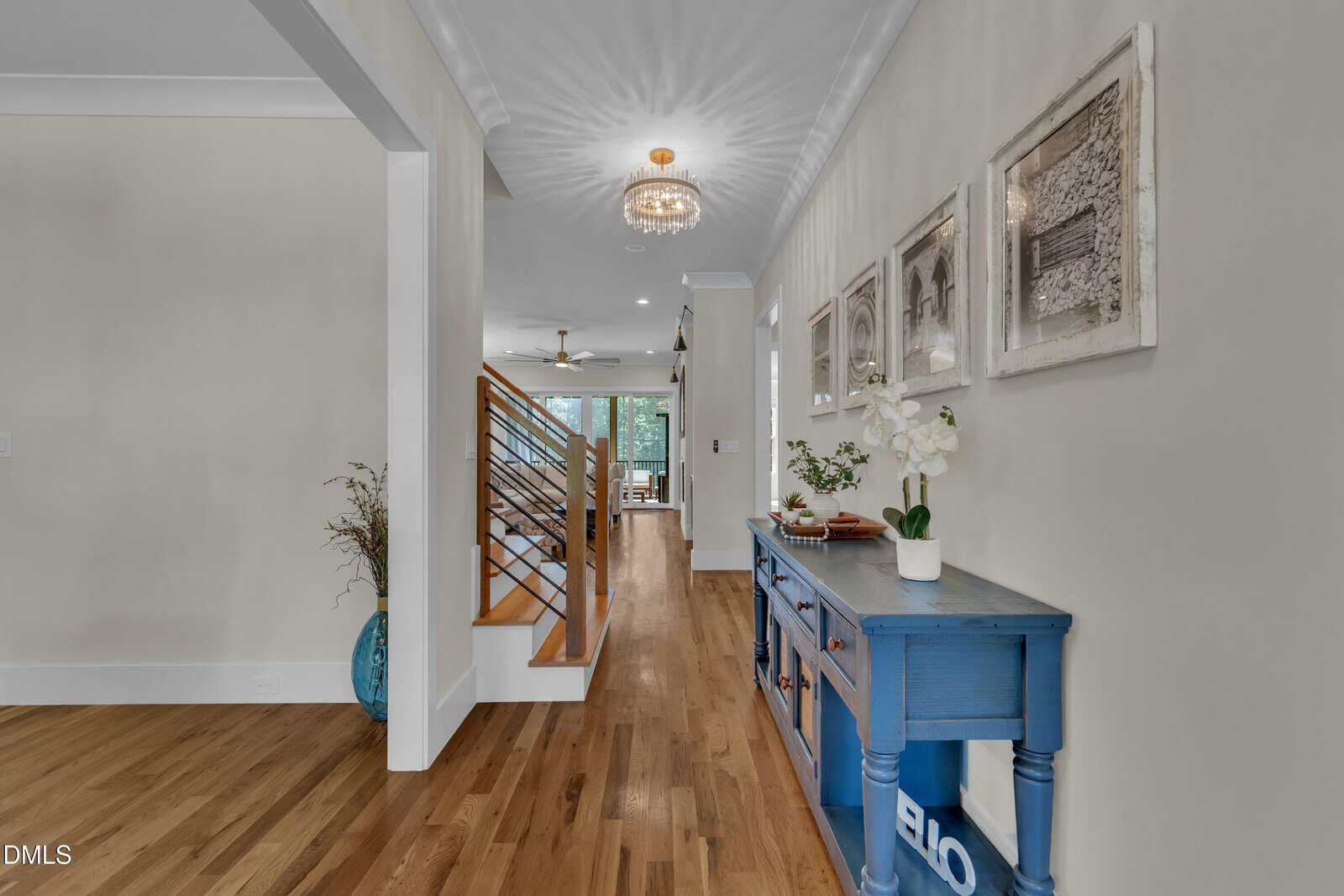 A hallway with light walls, hardwood floors, and a blue console table. Three framed pictures hang above the table. A modern chandelier hangs from the ceiling. Stairs with a wooden railing are on the left.