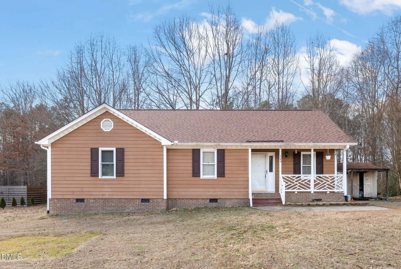 Single-story house with tan siding, brown shutters, a covered front porch, and a brick foundation located at 106 N Valley Drive in Louisburg. Leafless trees are in the background. The yard is mostly bare with patches of grass. A small shed is visible to the right.