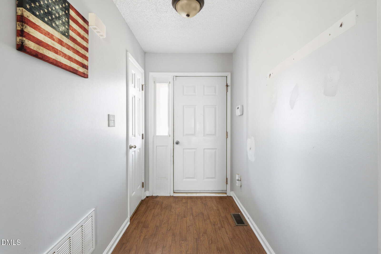A bright entryway with white doors, light gray walls, wood flooring, an American flag wall art, and visible marks on the right wall where items were removed.