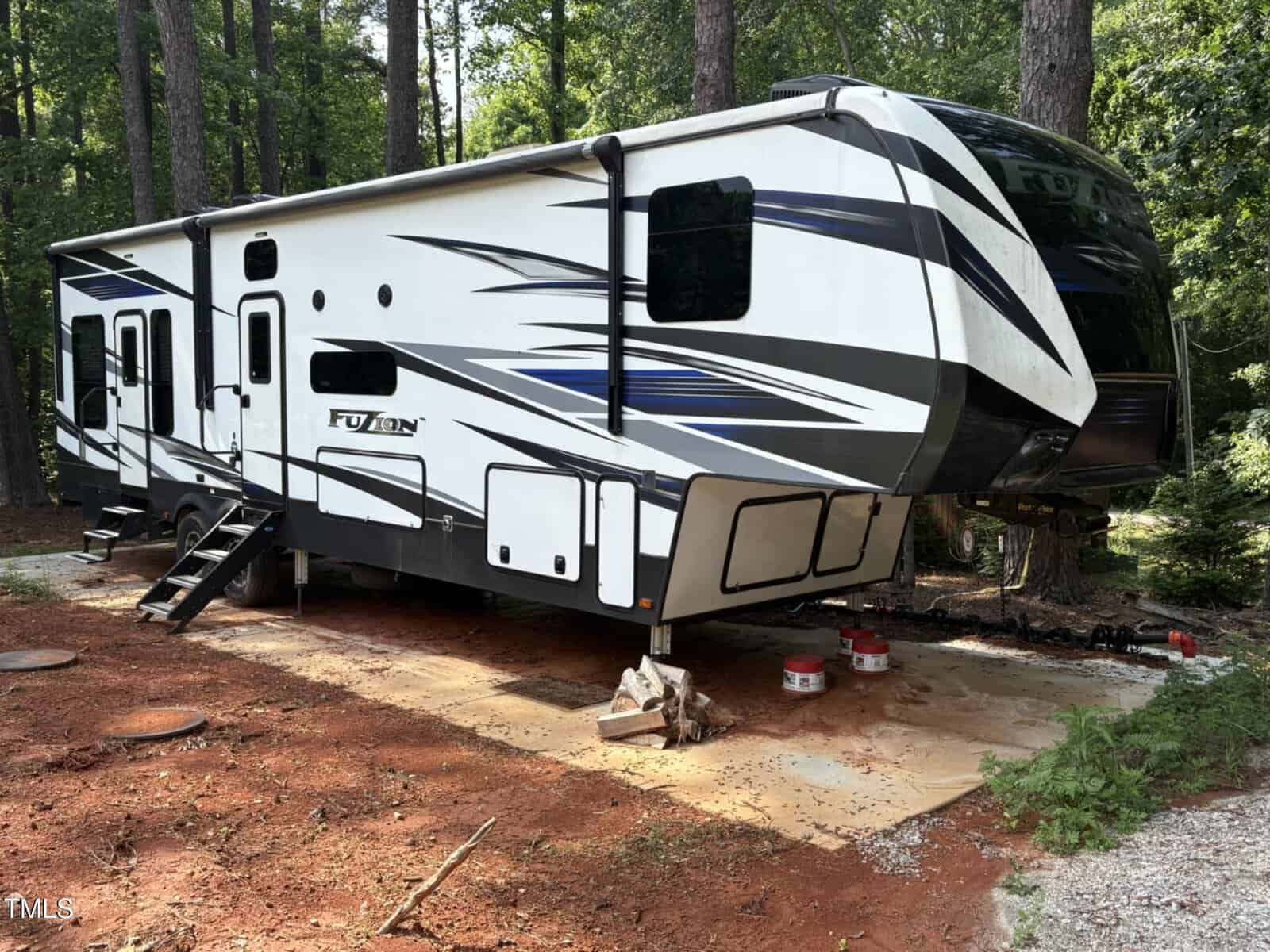 A large white and black fifth-wheel RV with blue accents is parked on a concrete pad at 147 Osage Drive, Louisburg, surrounded by tall trees. Steps lead to the entrance, and a few items are placed on the ground nearby.