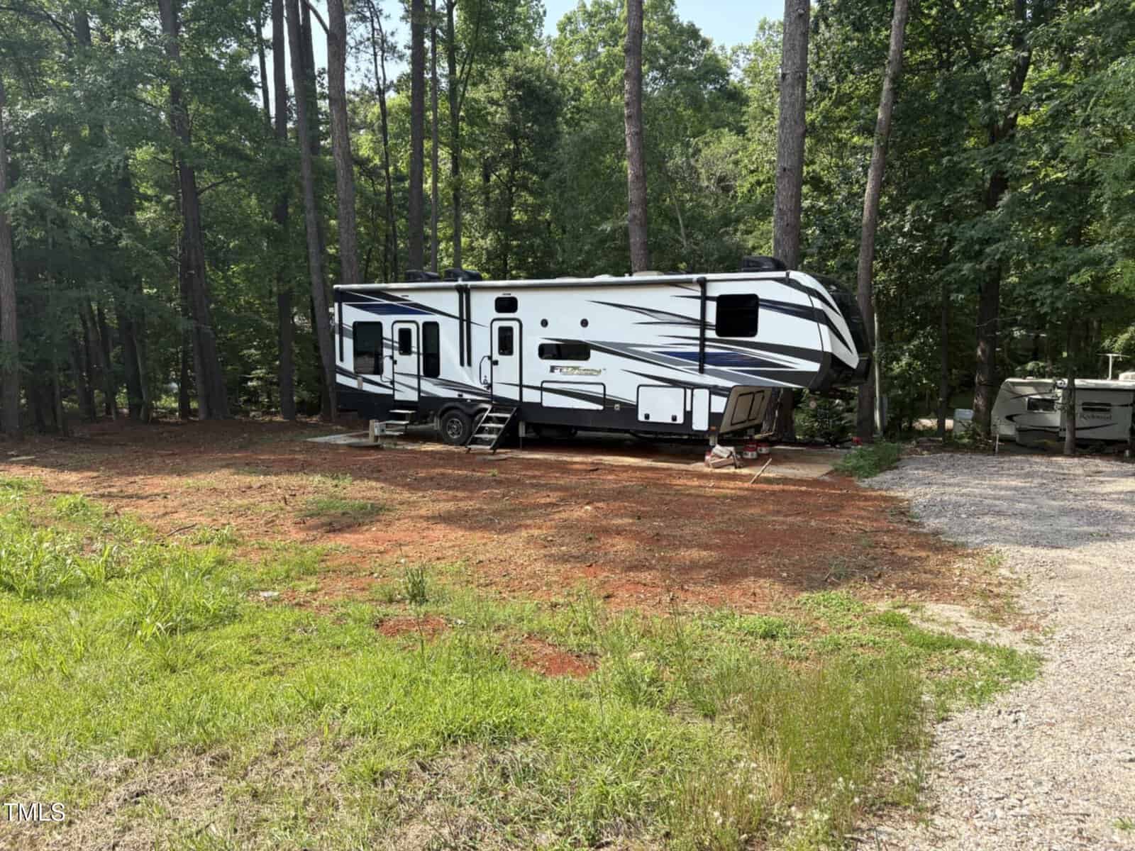 A white and black RV trailer is parked on a concrete pad in a wooded area with tall trees and sparse grass, adjacent to a gravel driveway.