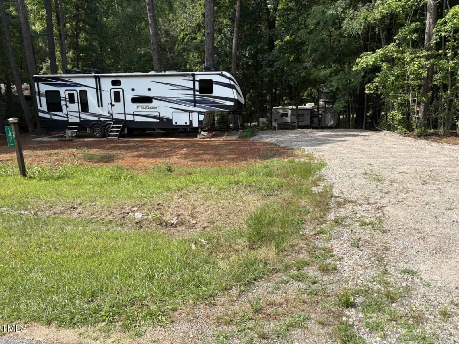 A white and black RV is parked on a dirt area near a gravel driveway surrounded by green grass and tall trees, with another RV visible in the background among the woods.