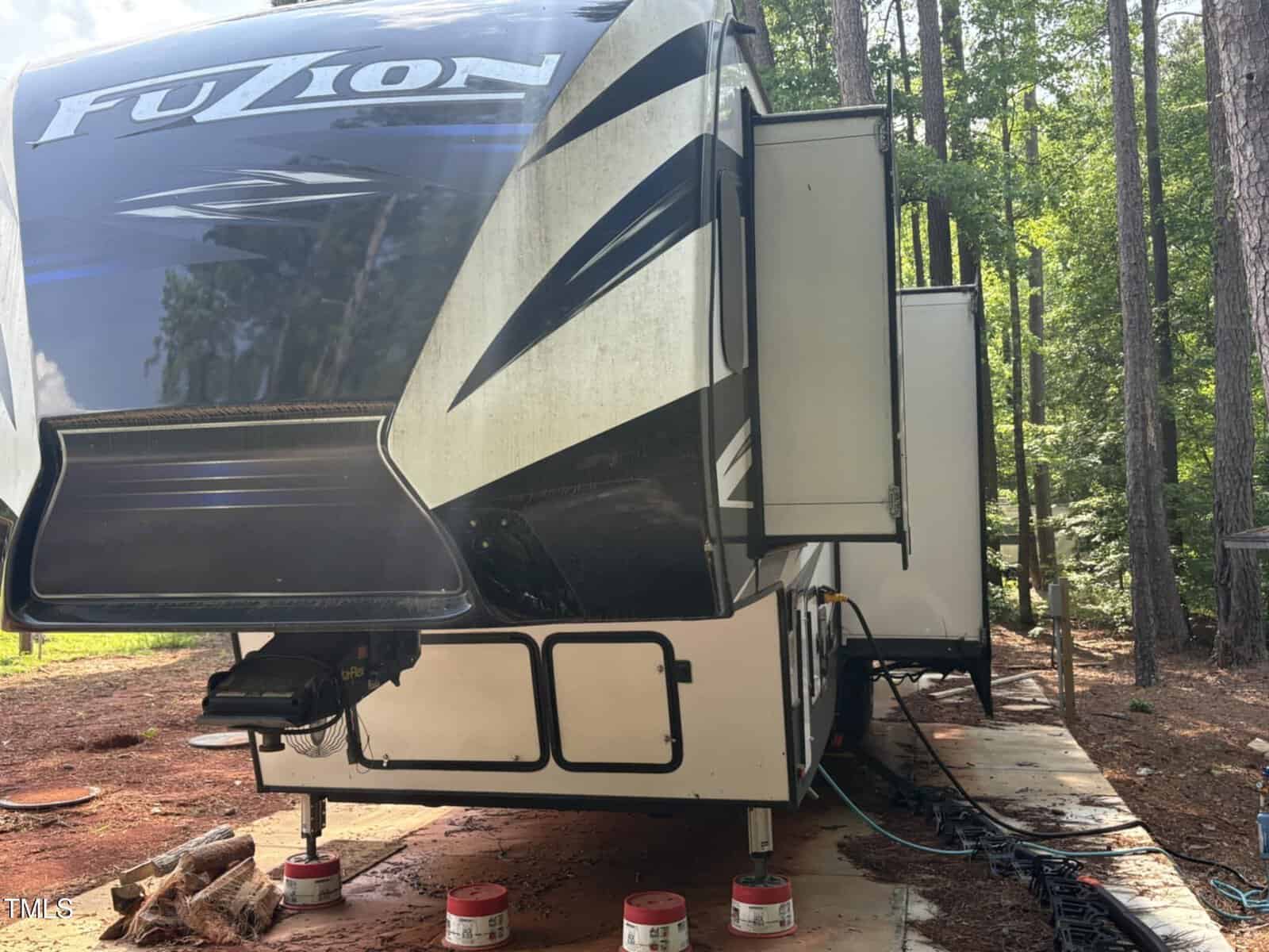 A large black and white RV labeled "Fuzion" is parked on a dirt lot surrounded by trees, with slide-outs extended and leveling jacks resting on red blocks. Cables and hoses are visible near the ground.