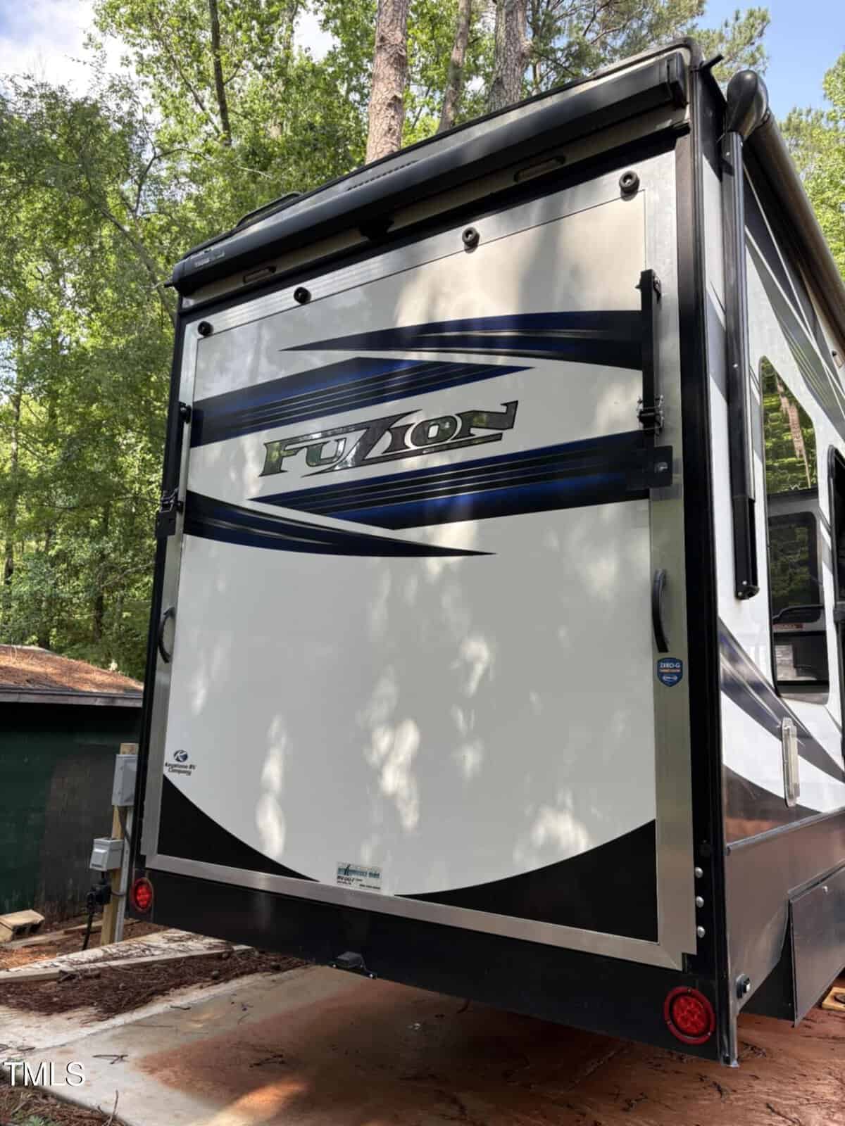 The rear view of a white RV with blue and black stripes, displaying the word "FUZION" on the back door. The RV is parked outdoors near trees and a wooden structure.